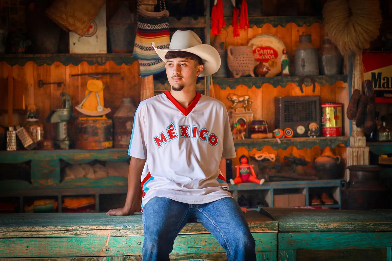 Young man wearing a Mexican jersey and cowboy hat in a rustic setting.